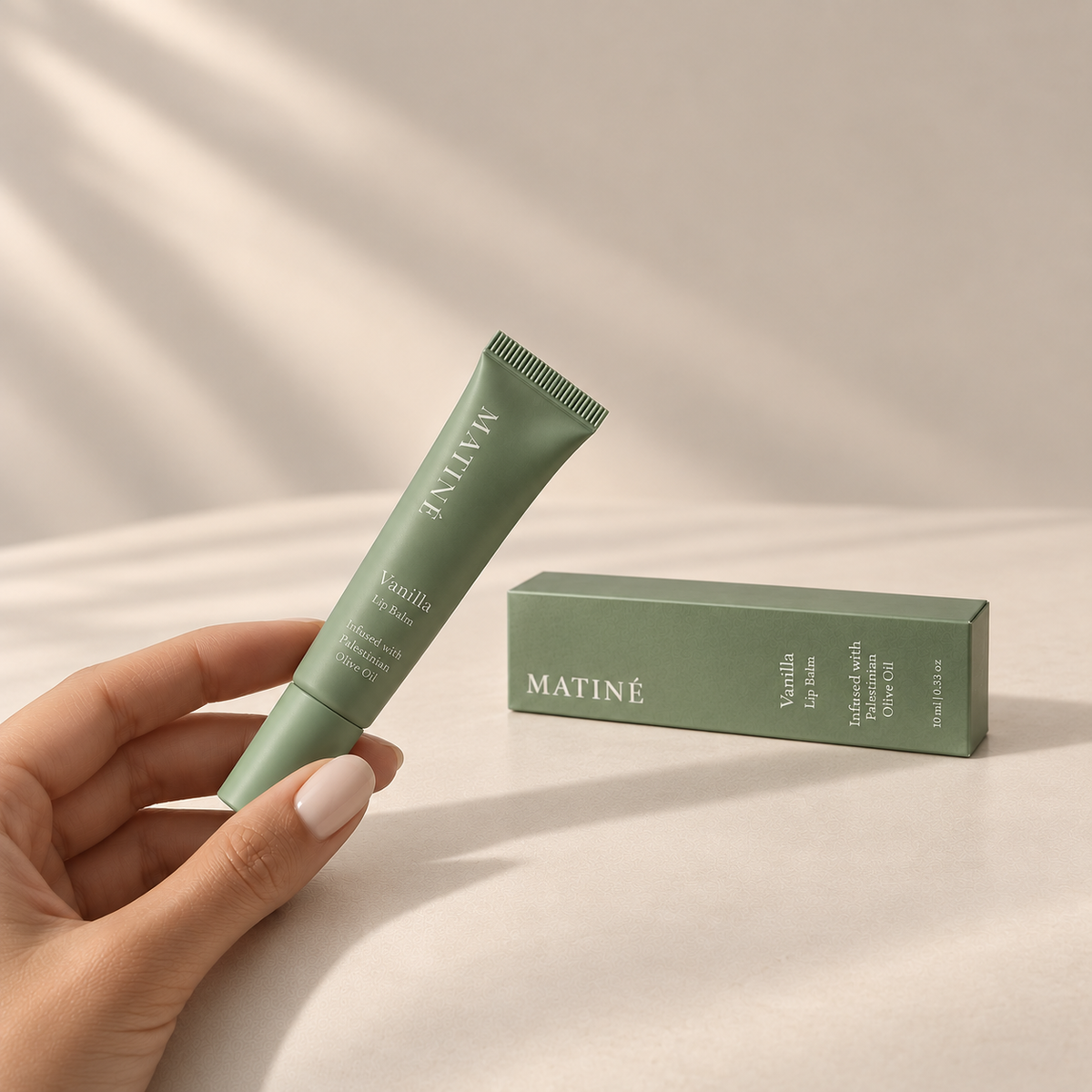 MATINÉ Vanilla Lip Balm held in hand beside its sage green box on a warm beige background.